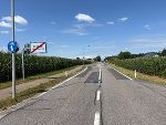 Bildbeschreibung: Landstraße in ländlicher Umgebung. Im Hintergrund ein Ortsschild.